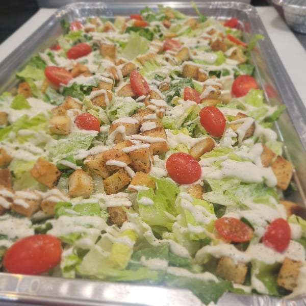 Caesar salad with romaine lettuce, cherry tomatoes, croutons, and creamy dressing in a metal tray.