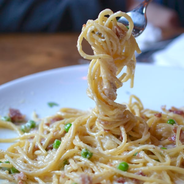 Fork lifting creamy spaghetti with peas and bacon from a white plate.