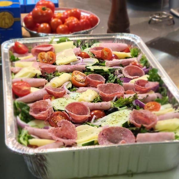 Large catering tray of antipasto salad with salami, ham, cheese, tomatoes, and cucumbers.