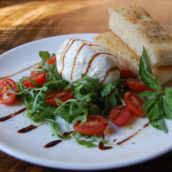 Burrata cheese with balsamic glaze, cherry tomatoes, arugula, basil, and focaccia bread on a white plate.