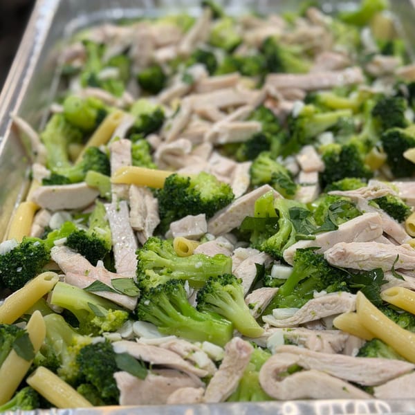 Close-up of penne pasta with chicken, broccoli, and herbs in a metal tray.