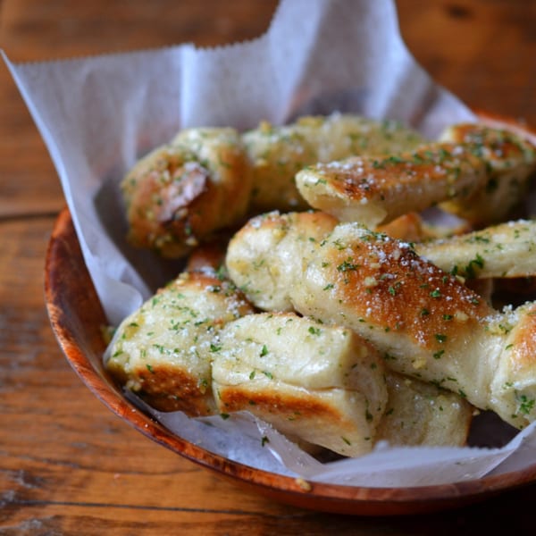 A wooden bowl filled with golden-brown garlic knots, sprinkled with parmesan cheese and parsley.
