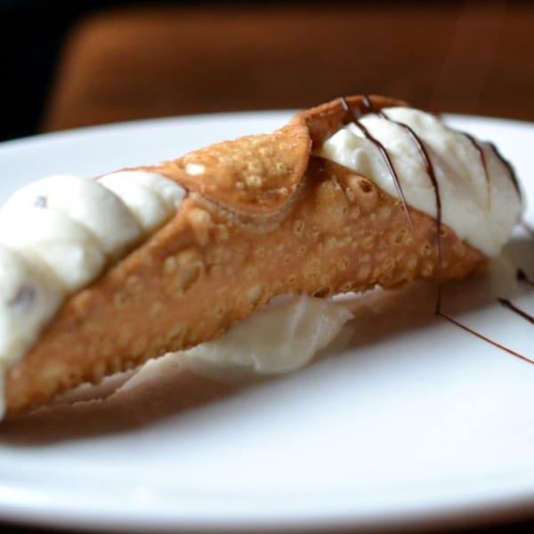 A cannoli from Danny's Pizzeria, filled with white cream and drizzled with chocolate, on a white plate.