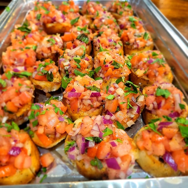 Close-up of many bruschetta appetizers with diced tomatoes, red onions, and parsley on a metal tray.