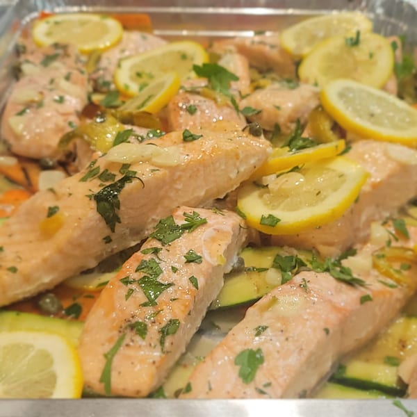 Baked salmon fillets with lemon slices, zucchini, and parsley in a silver pan.
