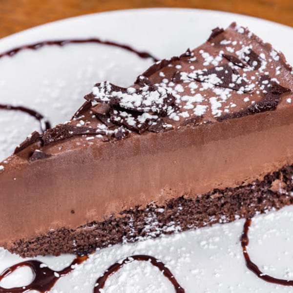 A slice of rich chocolate cake with powdered sugar and chocolate shavings on a white plate.