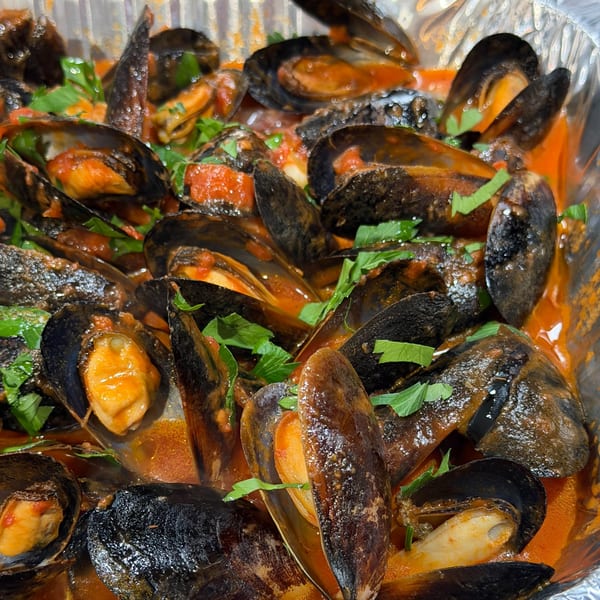 Steamed mussels in a rich red sauce with fresh parsley, served in a foil container.