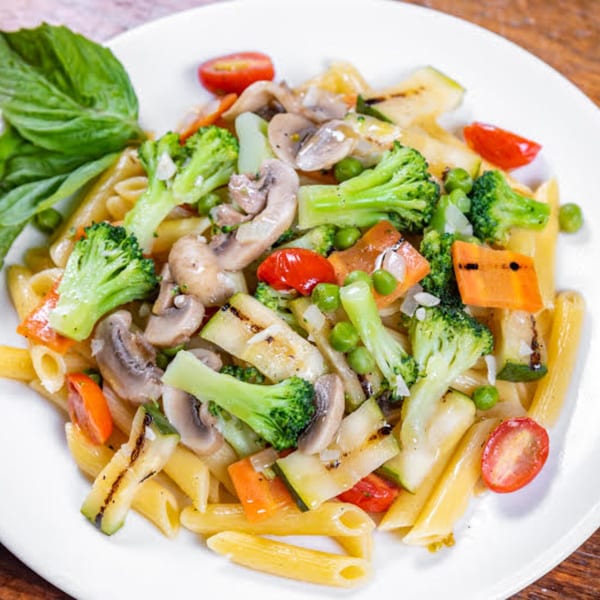 Penne pasta with broccoli, mushrooms, zucchini, peas, carrots, and cherry tomatoes, garnished with basil.