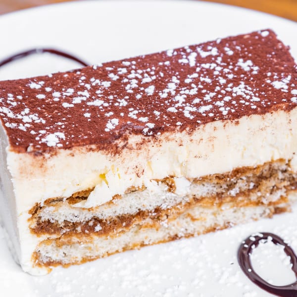 Close-up of a tiramisu slice with cocoa powder and powdered sugar on a white plate.